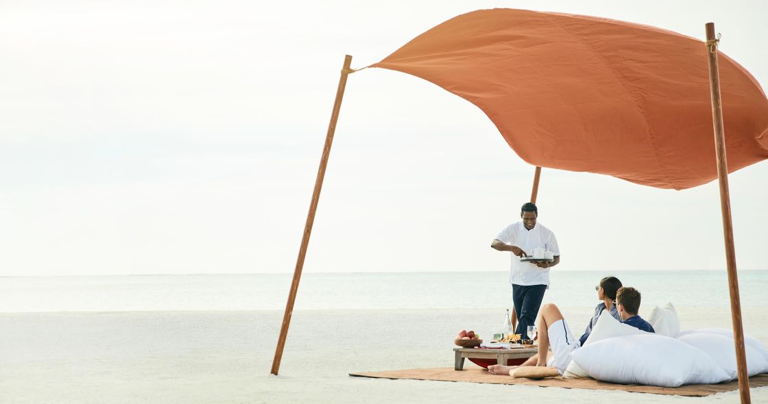 A Man Standing Next To A Couple Of Women Sitting Under An Umbrella