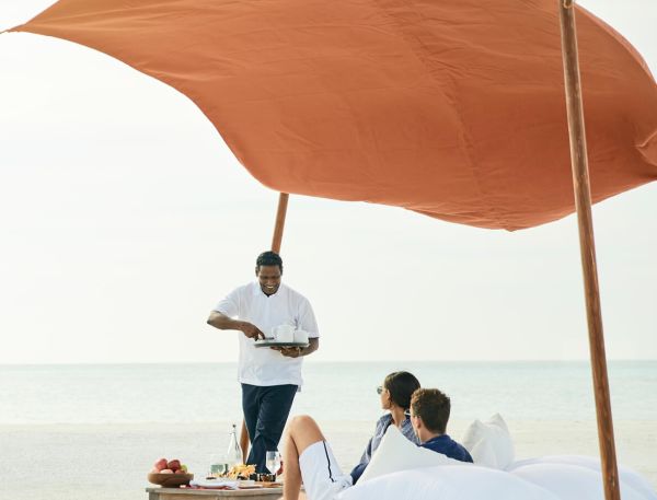 A Man Standing Next To A Couple Of Women Sitting Under An Umbrella